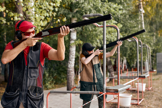 Side View On Couple Came in Shooting Range, Shooting As Hobby. Man And Woman shoot at metal flags, targets. Firearms pump-action shotgun. in goggles, headphones. glade with protection for shooting.