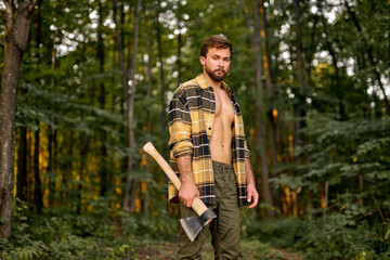 Logging season. Young Shirtless Caucasian Lumberman carry axe in forest, sexy handsome woodman....