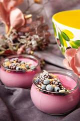Fresh summer vegan tasty violet creamy dessert with organic blackberries and almonds in a jar on the table  with pink pastel flowers and yellow bowl in the background. Food concept idea.
