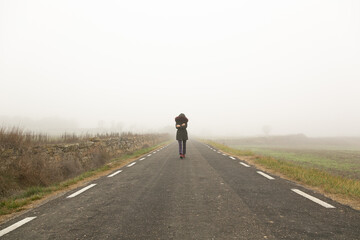 Chica con frío con abrigo caminando con niebla por una carretera secundaria,  huida 