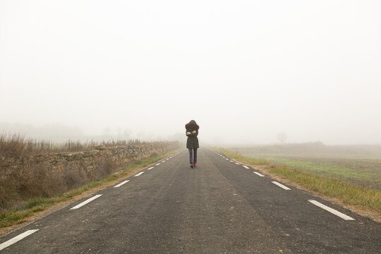 Chica con fr&iacute;o con abrigo caminando con niebla por una carretera secundaria,  huida 