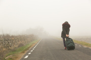Mujer abrigada en una carretera con niebla huyendo con una bolsa de maleta en solitario, refugiada