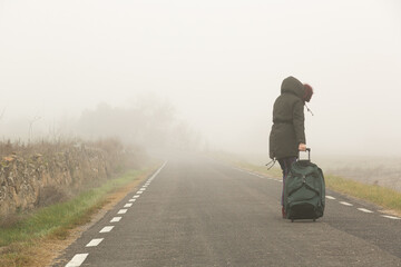 Mujer abrigada en una carretera con niebla huyendo con una bolsa de maleta en solitario, refugiada