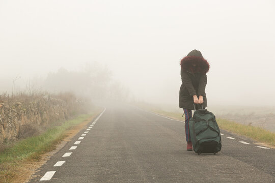 Mujer abrigada en una carretera con niebla huyendo con una bolsa de maleta en solitario, refugiada