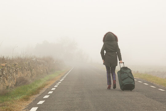 Con una bolsa maleta una mujer camina por la carretera con mucha niebla, parte trasera