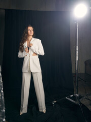 Confident young woman model in stylish white office pantsuit and high heel shoes poses for camera against black curtain in studio