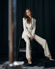 Long haired businesswoman in elegant formal white pantsuit with high heeled shoes sits on bar stool...