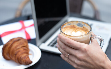 Hand's of young girl holds cup of coffee. On table laptop, present and plate of croissant. Work online, freelancer style modern business woman. outdoor/outside
