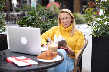 Happy girl sits in cafe have breakfast. On table coffee, croissant and laptop. Smile blonde in yellow sweater hold diary. Work online, freelancer style modern business woman. outdoor