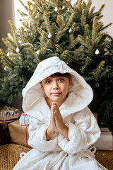 Obraz premium American Child Making Magic Wish for Winter Holidays. Boy Near the Christmas Tree at Home. Teen Boy In Bathrobe Praying. Waiting for Santa at Night. Holidays, Weekends, New Year, Christmas 2022