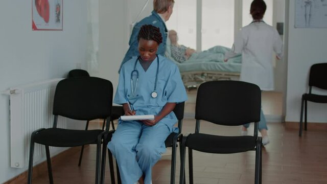 Portrait of african american nurse analyzing checkup papers, preparing for consultation in waiting area. Medical assistant looking at examination files, sitting in waiting room.