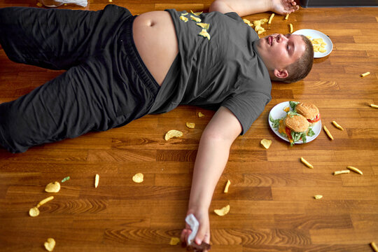 Overweight boy sleeping on floor surrounded by fast food, overeating. obese caucasian teenage boy in casual wear is lazy, spend rest time after school alone. healthcare, weight loss concept