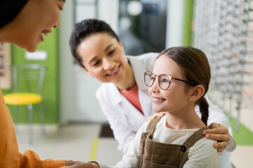 happy girl in eyeglasses smiling near asian mother and blurred oculist in optics store.