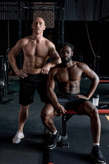 Break after hard crossfit training. Portrait of two serious sporty men having rest in gym after workout. Diverse mixed-race confident male fitness models posing at camera, having strong muscular body