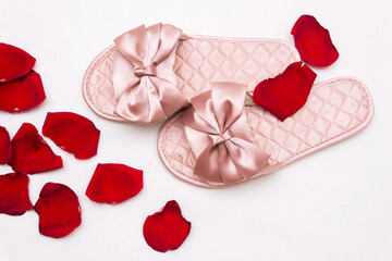 Close-up of pink satin female glamorous stylish home slippers with bows with red rose petals in the foreground isolated on a white background. Top view