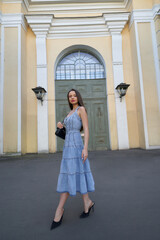 Fototapeta na wymiar Stylish brunette woman wearing long denim summer dress holds cup of drink standing near building with vintage door low angle shot