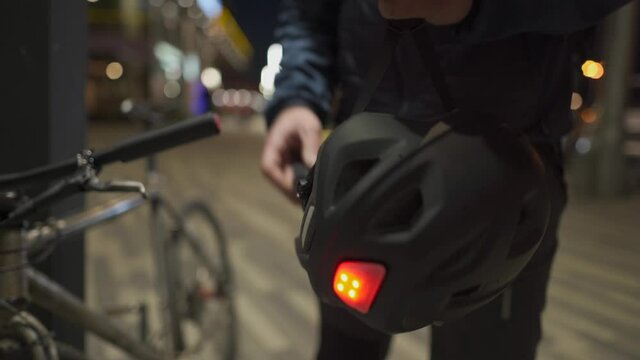 Male cyclist in protective medical mask against air pollution, smog, viruses, infection and allergies puts on bicycle helmet with red lamp at night in city takes bike from parking lot after work.