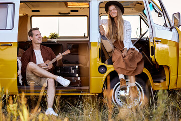 Young hipster couple sitting in camper van playing ukulele, making music together. Adorable female in hat and handsome guy relaxing, having rest together, romantic time during adventure, travel - Powered by Adobe