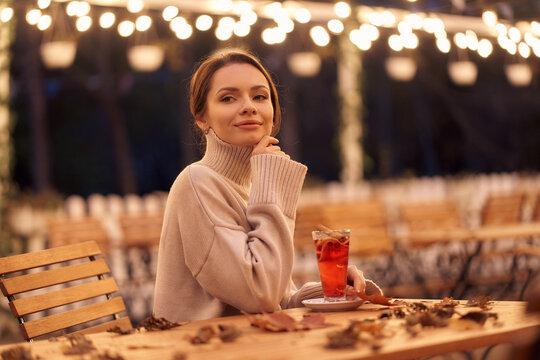 Thoughtful woman in knitted clothes sits at wooden table with mulled wine glass and scattered dry leaves on outdoor terrace in evening