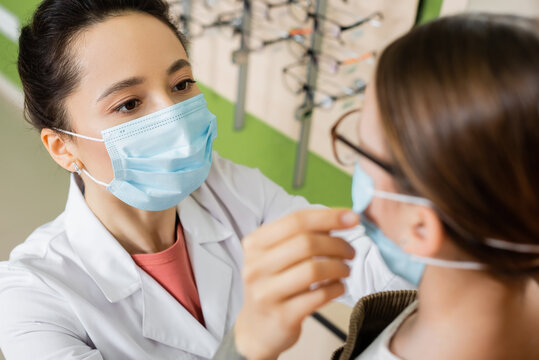 Oculist In Medical Mask Trying Eyeglasses On Blurred Girl In Optics Store.