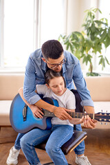 Fototapeta premium Handsome man and cute child girl playing song on guitar instrument showing sounds sitting on sofa in house living room enjoying daydream together, side view. caucasian adult and child perform music