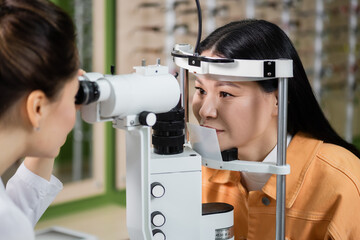 blurred optometrist testing eyesight of asian woman on vision screener in optics shop.