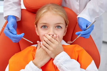 Top view on caucasian frightened child girl covering mouth by hand while treating with dental tools for oral check-up held by cropped professional dentist in blue gloves. medical healthcare