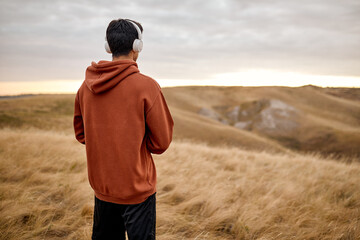 View from back on unrecognizable brunette male in casual red hoodie looking forward at empty field, keep calm, meditating with calm music in headphones, after training workout