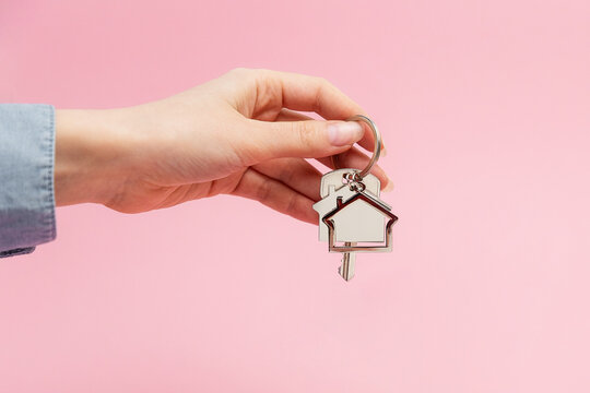 Female Hand Holding Key And House Keychain. Pink Background With Copy Space. Concept Of Mortgage And Leasing Of Real Estate