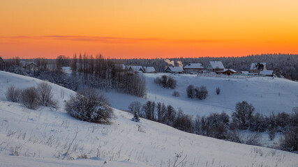 View of hills covered in snow with sparse trees and a small village against an orange sky.  Travel concept