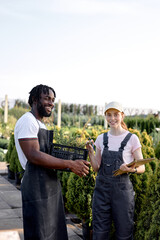 team of researchers at work in greenhouse, female with notepad and pen in hand, stand talking with black guy in uniform. Greenhouse production. Female farm worker examine and checking plants