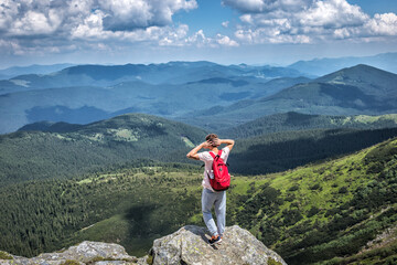 active tourist hiker with a backpack on top of a mountain in summer stands at a height and looks at the green valley