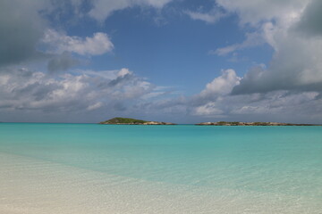 Fototapeta premium Exuma Bahamas white sand beach with blue skies, white clouds, small cays in the background. Blue green, turquoise, and relaxing water invokes calm during vacation. Deserted, secluded romantic beach.