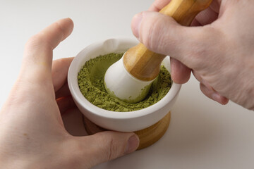 The matcha is rubbed in a white bowl with a pestle. Male hands are rubbing green powder in a mortar.