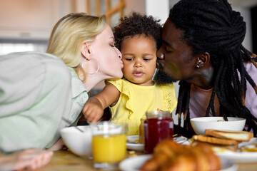 Loving parents kissing child girl sitting behind table during breakfast or lunch. beautiful family spend weekends together, enjoy having meal in cozy kitchen at home. delicious food, love concept