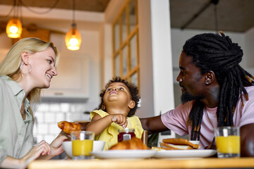 happy caucasian family of man and woman sitting together talking with child girl while eating healthy breakfast at home, in cozy bright room. excited family have fun while having meal
