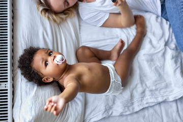 Obraz na płótnie Canvas top view on cute child girl with black skin lying on bed with mother, with baby pacifier in mouth. view from above on shirtless kid after waking up in the morning. children, lifestyle concept