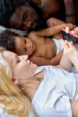 Obraz na płótnie Canvas father mother and kid girl in diaper playing smartphone on bed in bedroom. Loving diverse Parents and little black child are resting at home. View from above. leisure, weekends. focus on child