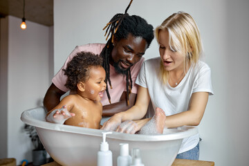 Nice black man and blonde female washing daughter in bath, have fun, talking, explaining, playing with toys. in cozy bright room at home. young family with diverse appearance enjoy time together