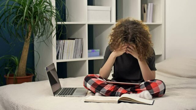 Difficulties of distance learning. Teenage girl doing homework sitting on the bed at home. Not everything works out, the girl is upset.