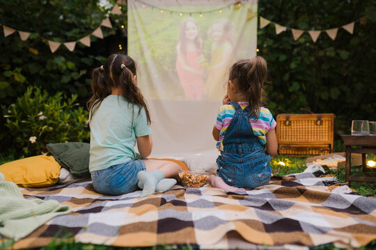 Backyard Family Outdoor Movie Night With Kids. Sisters Spending Time Together And Watching Cimema At Backyard. DIY Screen With Film. Summer Outdoor Weekend Activities With Children. Open Air Cinema.