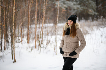 Fototapeta na wymiar Portrait of a red-haired girl in winter forest in warm clothes: black hat and light jacket looking at the camera. Blurred forest background