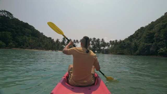 Man with sunglasses and hat rows pink plastic canoe along sea against green hilly islands with wild jungles. Traveling to tropical countries.