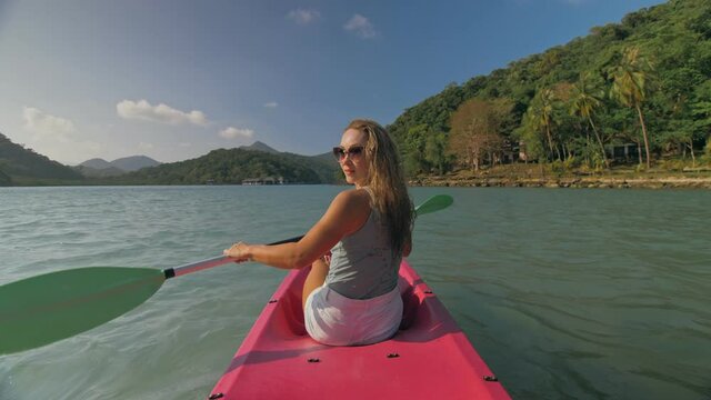 Long haired blonde woman with sunglasses rows bright pink canoe along sea bay water to beach with growing palms.