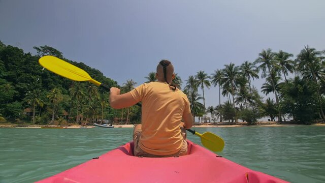 Man with sunglasses and hat rows pink plastic canoe along sea against green hilly islands with wild jungles. Traveling to tropical countries.