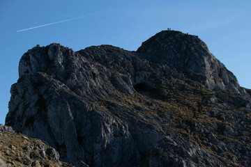 Cumbre del Udalatx en el Pais Vasco, con personas en la cima y la cueva de Mari bajo esta en un día de cielo azul.
