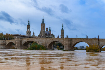 Obraz premium Puente de Piedra bridge over Ebro river with yellow water against the backdrop of the towers of Cathedral-Basilica of Our Lady of the Pillar in Zaragoza, Spain. Autumn cityscape of spanish city