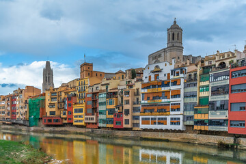 Colorful bright houses of Girona old town are reflected in the water of the Onyar river, Spain. Cityscape of spanish tourist city with two towers against blue cloudy sky