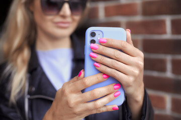 Blonde stylish woman make photo on mobile phone. Female hands with bright neon pink manicure hold a smartphone in a purple case
