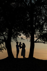 silhouettes of a couple in love , holding a child in their hands , stand between the trees and look at the sea